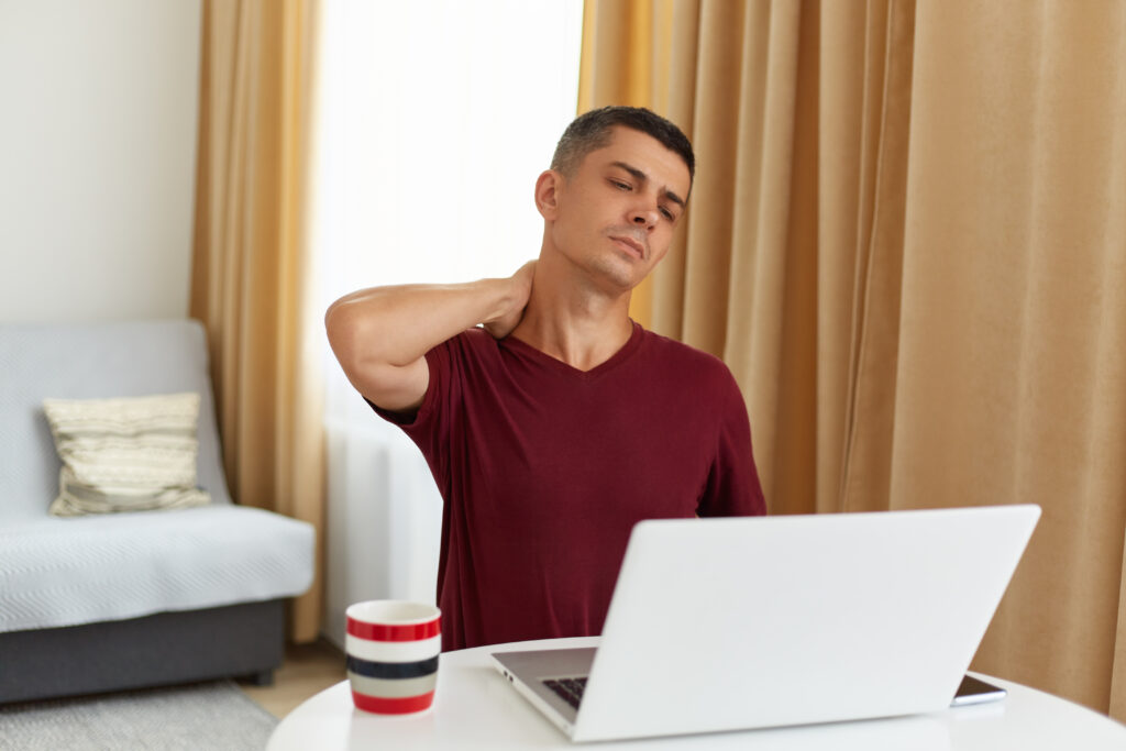 indoor shot of tired man working online at home, sitting at table in living room against sofa, having lots freelance work, having pain in neck, massaging, looking at laptop display.